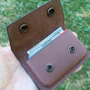 Puede incluir: Una cartera de cuero marr&oacute;n con cierre de solapa asegurado por dos botones de presi&oacute;n de metal de lat&oacute;n antiguo. El compartimento interior contiene un billete de cien d&oacute;lares estadounidense doblado. La cartera tiene costuras visibles.
