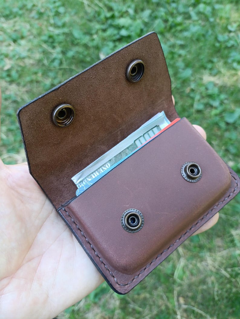 Puede incluir: Una cartera de cuero marr&oacute;n con cierre de solapa asegurado por dos botones de presi&oacute;n de metal de lat&oacute;n antiguo. El compartimento interior contiene un billete de cien d&oacute;lares estadounidense doblado. La cartera tiene costuras visibles.