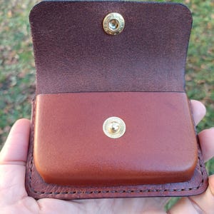 May include: A brown leather pouch with a flap closure featuring a gold-toned metal snap button. The interior of the flap shows the natural rough texture of the leather, while the main body of the pouch has a smooth, finished surface.