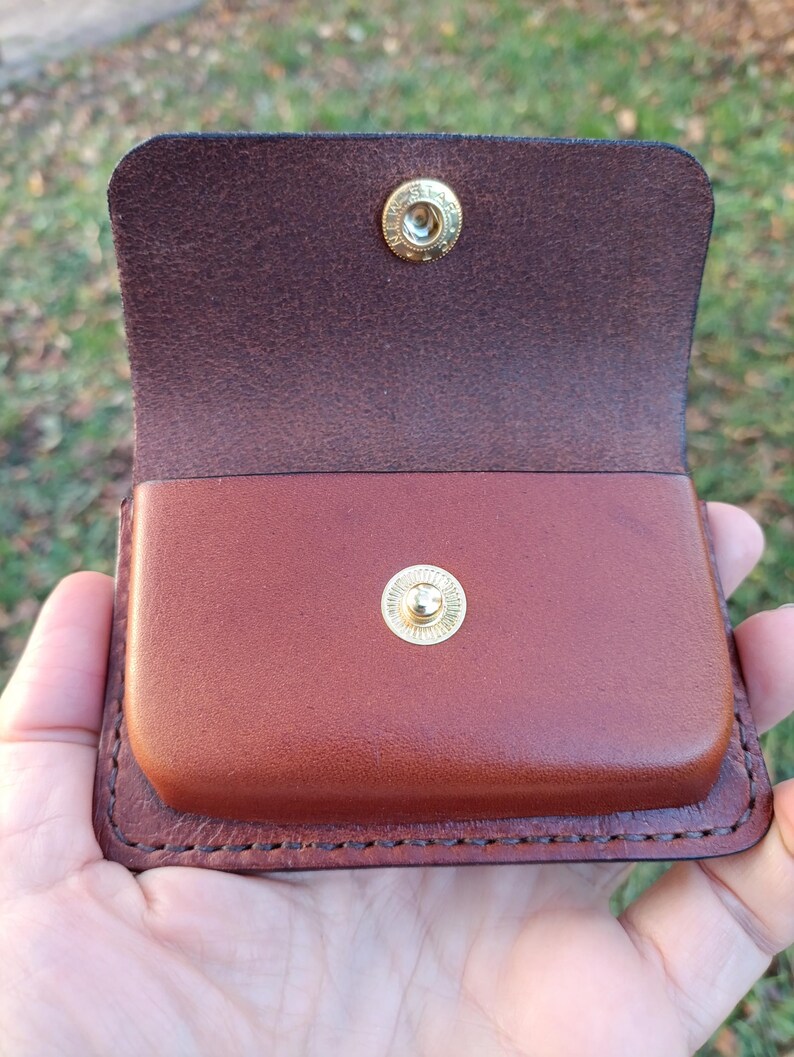 May include: A brown leather pouch with a flap closure featuring a gold-toned metal snap button. The interior of the flap shows the natural rough texture of the leather, while the main body of the pouch has a smooth, finished surface.