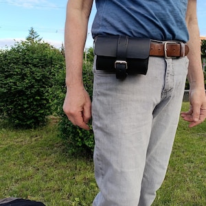 May include: A black leather belt pouch with a buckle closure, attached to a brown leather belt. The pouch is worn on the waist over gray jeans and a blue shirt. The background shows green bushes and a blue sky.