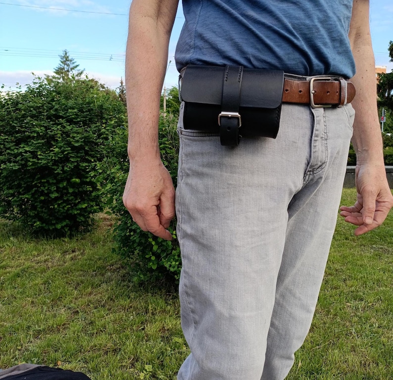 May include: A black leather belt pouch with a buckle closure, attached to a brown leather belt. The pouch is worn on the waist over gray jeans and a blue shirt. The background shows green bushes and a blue sky.