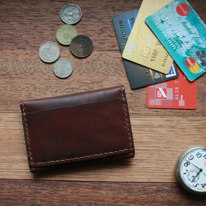May include: A brown leather wallet, credit cards, coins, and a silver pocket watch with a white face are arranged on a wooden surface. The wallet is rectangular with visible stitching. The watch has a chain.