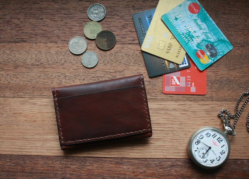 May include: A brown leather wallet, credit cards, coins, and a silver pocket watch with a white face are arranged on a wooden surface. The wallet is rectangular with visible stitching. The watch has a chain.
