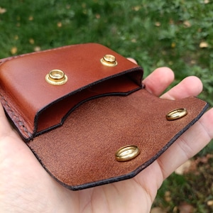 Puede incluir: Una peque&ntilde;a bolsa de cuero marr&oacute;n con un exterior liso y un interior texturizado. La bolsa se asegura con dos broches de presi&oacute;n de metal dorado en la solapa y dos broches de presi&oacute;n correspondientes en el cuerpo principal.