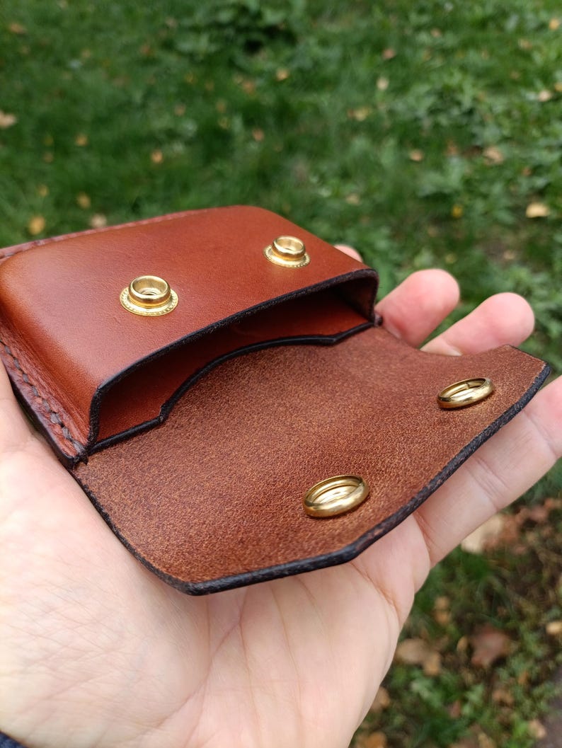 Puede incluir: Una peque&ntilde;a bolsa de cuero marr&oacute;n con un exterior liso y un interior texturizado. La bolsa se asegura con dos broches de presi&oacute;n de metal dorado en la solapa y dos broches de presi&oacute;n correspondientes en el cuerpo principal.