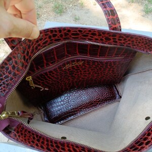 May include: A deep red tote bag with a textured crocodile-embossed pattern. The interior features a light beige lining, a zippered pocket with gold-toned hardware, and a gold-colored brand logo printed on the inner pocket panel.