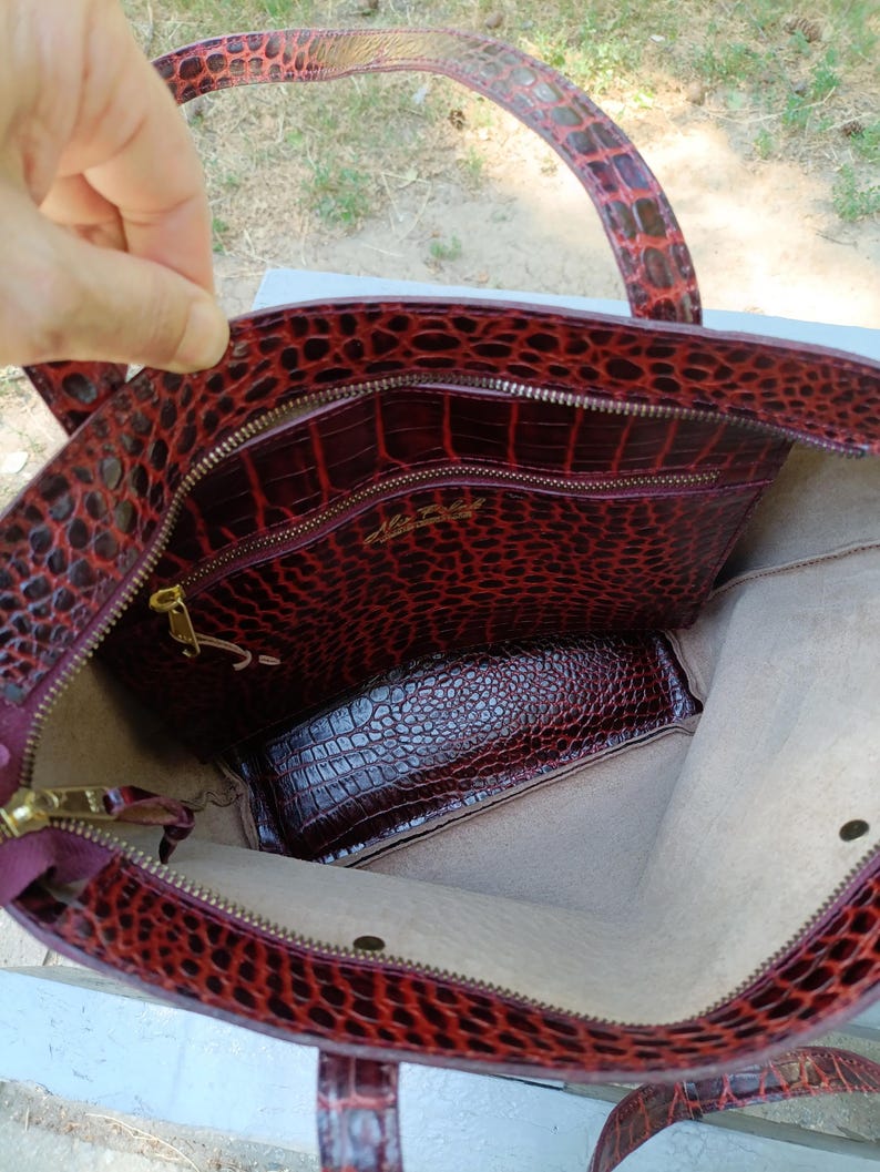 May include: A deep red tote bag with a textured crocodile-embossed pattern. The interior features a light beige lining, a zippered pocket with gold-toned hardware, and a gold-colored brand logo printed on the inner pocket panel.