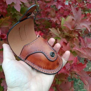 K&ouml;nnte beinhalten: Eine handgefertigte, braune Lederh&uuml;lle mit Schnappverschluss. Die H&uuml;lle hat ein geschwungenes Design mit dekorativen N&auml;hten und einem helleren braunen Innenraum. Der Hintergrund zeigt rotes und gr&uuml;nes Herbstlaub.