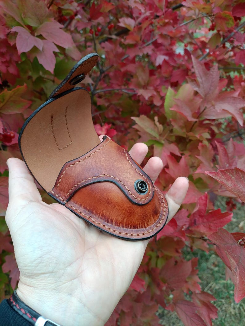 K&ouml;nnte beinhalten: Eine handgefertigte, braune Lederh&uuml;lle mit Schnappverschluss. Die H&uuml;lle hat ein geschwungenes Design mit dekorativen N&auml;hten und einem helleren braunen Innenraum. Der Hintergrund zeigt rotes und gr&uuml;nes Herbstlaub.