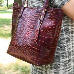 May include: A deep burgundy tote bag featuring a textured crocodile skin pattern. The bag has two vertical straps attached with gold-toned metal rivets at the top. The surface has a glossy finish with a dark red and black color gradient.