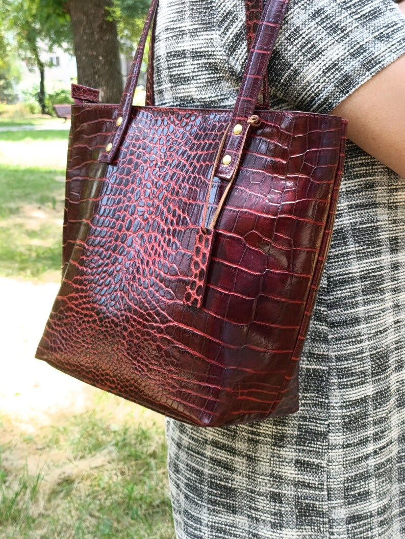 May include: A deep burgundy tote bag featuring a textured crocodile skin pattern. The bag has two vertical straps attached with gold-toned metal rivets at the top. The surface has a glossy finish with a dark red and black color gradient.