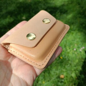 May include: A light tan leather wallet featuring a rectangular flap secured by two polished gold-tone metal snap buttons. The wallet has visible stitching along the edges and a smooth, natural finish on the exterior surface of the leather.