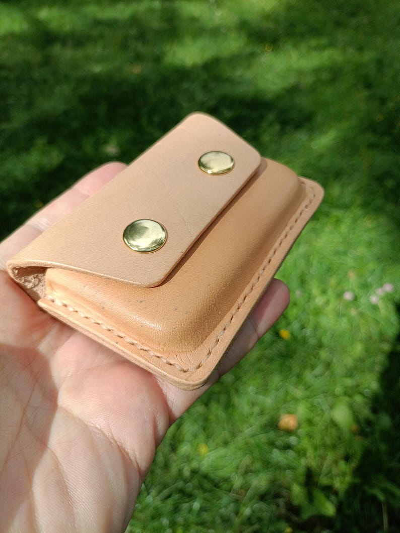 May include: A light tan leather wallet featuring a rectangular flap secured by two polished gold-tone metal snap buttons. The wallet has visible stitching along the edges and a smooth, natural finish on the exterior surface of the leather.