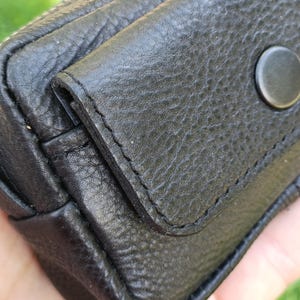 Puede incluir: Un estuche de cuero negro texturizado con cierre de solapa asegurado por un bot&oacute;n de presi&oacute;n circular negro mate. El exterior muestra costuras visibles a lo largo de los bordes de la solapa y del cuerpo principal del peque&ntilde;o contenedor.