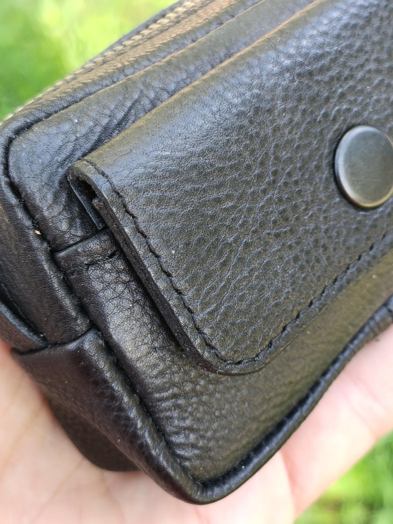 Puede incluir: Un estuche de cuero negro texturizado con cierre de solapa asegurado por un bot&oacute;n de presi&oacute;n circular negro mate. El exterior muestra costuras visibles a lo largo de los bordes de la solapa y del cuerpo principal del peque&ntilde;o contenedor.