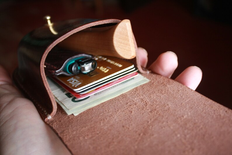 May include: A brown leather wallet with a wooden accent, open to reveal a lighter, credit cards, and folded bills. The wallet is held open, showcasing its contents. The leather has a natural, textured appearance.
