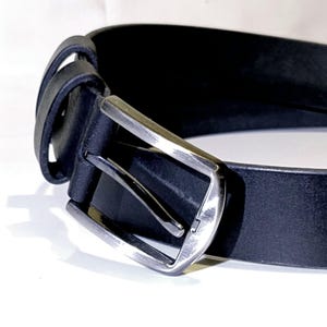 May include: A close-up shot of a black leather belt with a silver buckle. The belt features a classic design with a rectangular buckle and a single belt loop. The leather appears smooth, and the belt is set against a white backdrop.