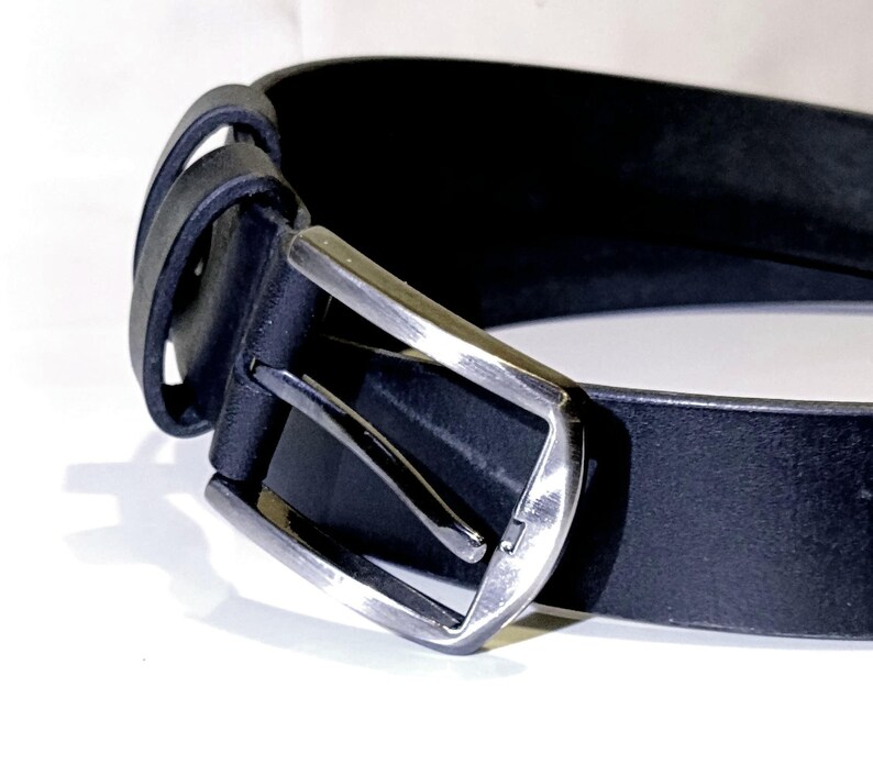 May include: A close-up shot of a black leather belt with a silver buckle. The belt features a classic design with a rectangular buckle and a single belt loop. The leather appears smooth, and the belt is set against a white backdrop.