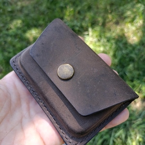 May include: A dark brown leather card holder with a rectangular shape and rounded corners. The accessory features a single flap closure secured by a centered, circular, antique brass-toned metal snap button. Visible stitching runs along the edges.