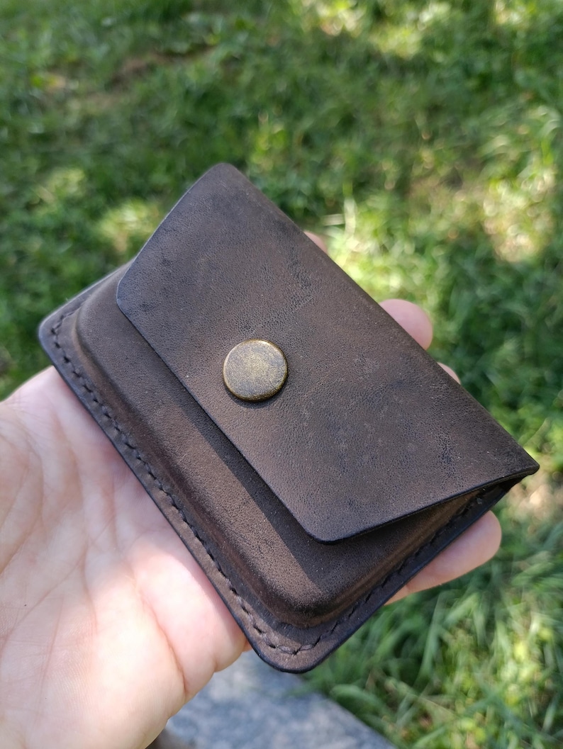 May include: A dark brown leather card holder with a rectangular shape and rounded corners. The accessory features a single flap closure secured by a centered, circular, antique brass-toned metal snap button. Visible stitching runs along the edges.