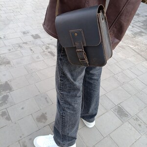 Pu&ograve; includere: Una borsa a tracolla in pelle marrone scuro con chiusura a fibbia e tracolla. La borsa &egrave; indossata sulla spalla di una persona che indossa jeans e una giacca di pelle marrone.
