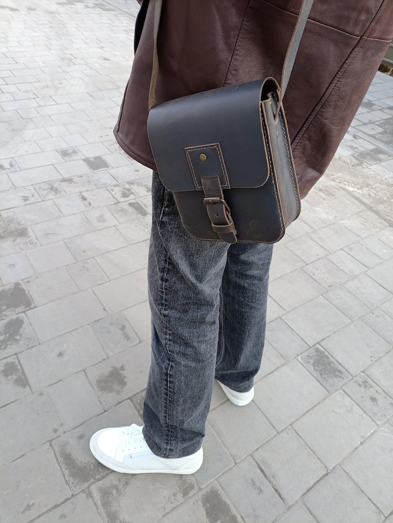 Pu&ograve; includere: Una borsa a tracolla in pelle marrone scuro con chiusura a fibbia e tracolla. La borsa &egrave; indossata sulla spalla di una persona che indossa jeans e una giacca di pelle marrone.