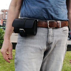 May include: A black leather belt pouch with a buckle closure is attached to a brown leather belt. The belt is worn with gray jeans and a blue t-shirt. The pouch is designed to be worn around the waist.