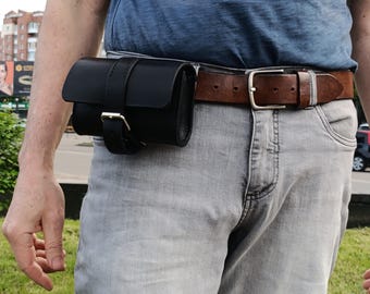 May include: A black leather belt pouch with a buckle closure is attached to a brown leather belt. The belt is worn with gray jeans and a blue t-shirt. The pouch is designed to be worn around the waist.