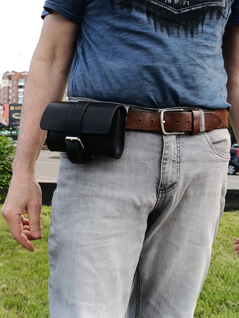 May include: A black leather belt pouch with a buckle closure is attached to a brown leather belt. The belt is worn with gray jeans and a blue t-shirt. The pouch is designed to be worn around the waist.