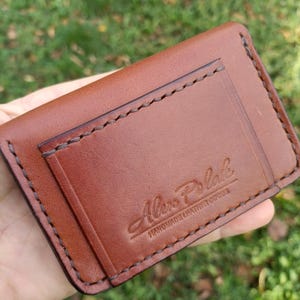 May include: A brown leather card holder featuring visible dark brown stitching along the edges and a pocket. The front is embossed with the text Alex Polak HANDMADE LEATHER GOODS in a script and block font combination.