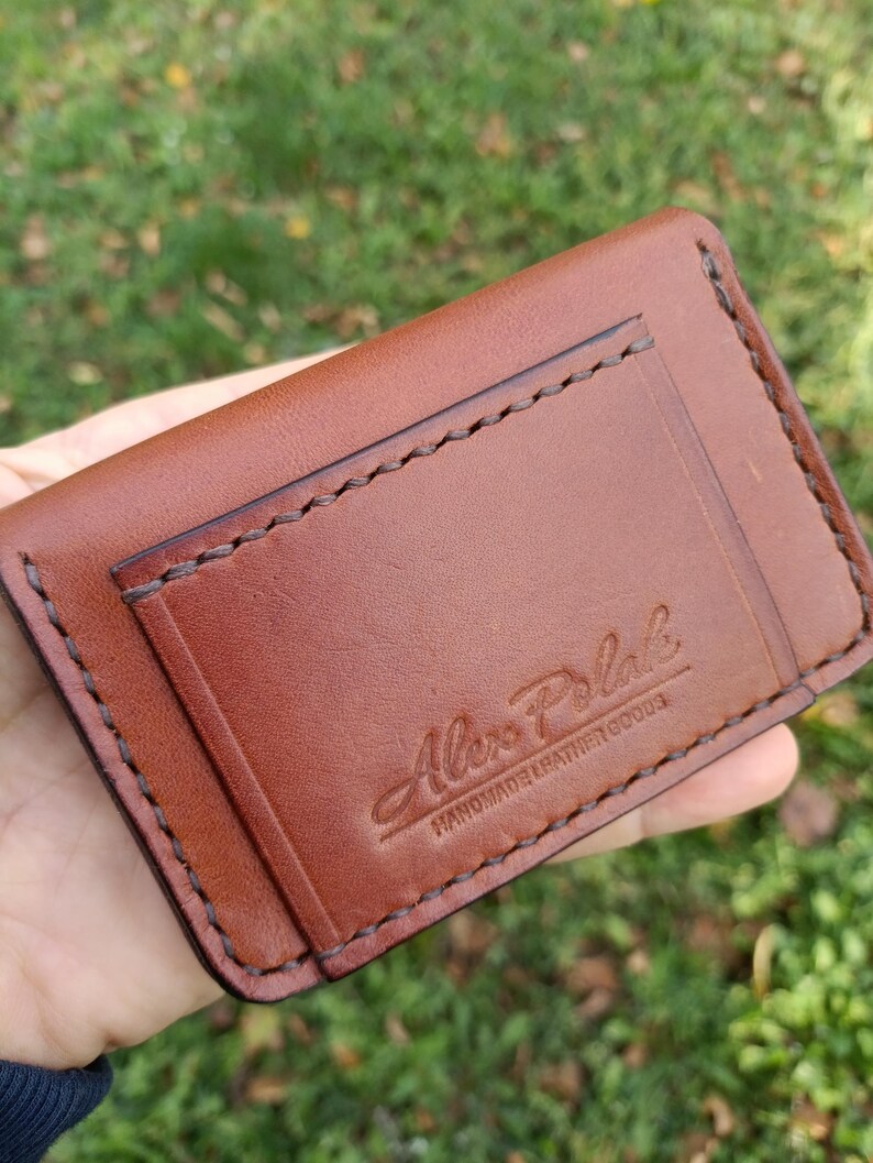 May include: A brown leather card holder featuring visible dark brown stitching along the edges and a pocket. The front is embossed with the text Alex Polak HANDMADE LEATHER GOODS in a script and block font combination.