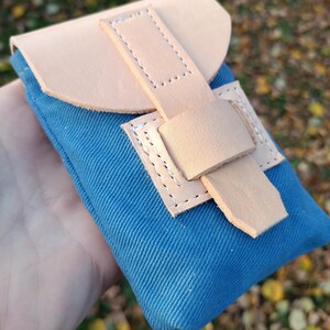 May include: A small blue canvas pouch featuring a light tan leather flap and a matching leather strap closure. The leather components are secured with visible white stitching, creating a contrast against the textured blue fabric body.