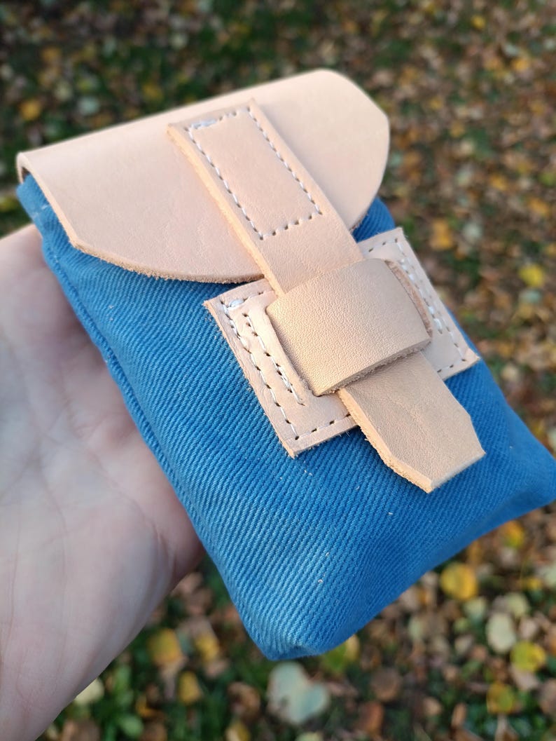 May include: A small blue canvas pouch featuring a light tan leather flap and a matching leather strap closure. The leather components are secured with visible white stitching, creating a contrast against the textured blue fabric body.