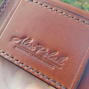 May include: A brown leather wallet featuring visible dark stitching along the edges. The surface is embossed with the text Alex Polak HANDMADE LEATHER GOODS in a combination of script and block lettering centred on the front panel.
