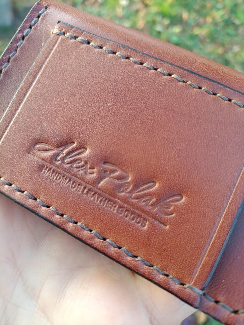 May include: A brown leather wallet featuring visible dark stitching along the edges. The surface is embossed with the text Alex Polak HANDMADE LEATHER GOODS in a combination of script and block lettering centred on the front panel.
