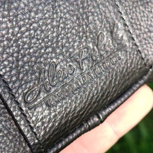 Puede incluir: Una cartera de cuero negro texturizado con el texto en relieve Alex Polak y las palabras HANDMADE LEATHER GOODS. La superficie tiene un patr&oacute;n de grano con costuras visibles a lo largo del borde del material de cuero doblado.