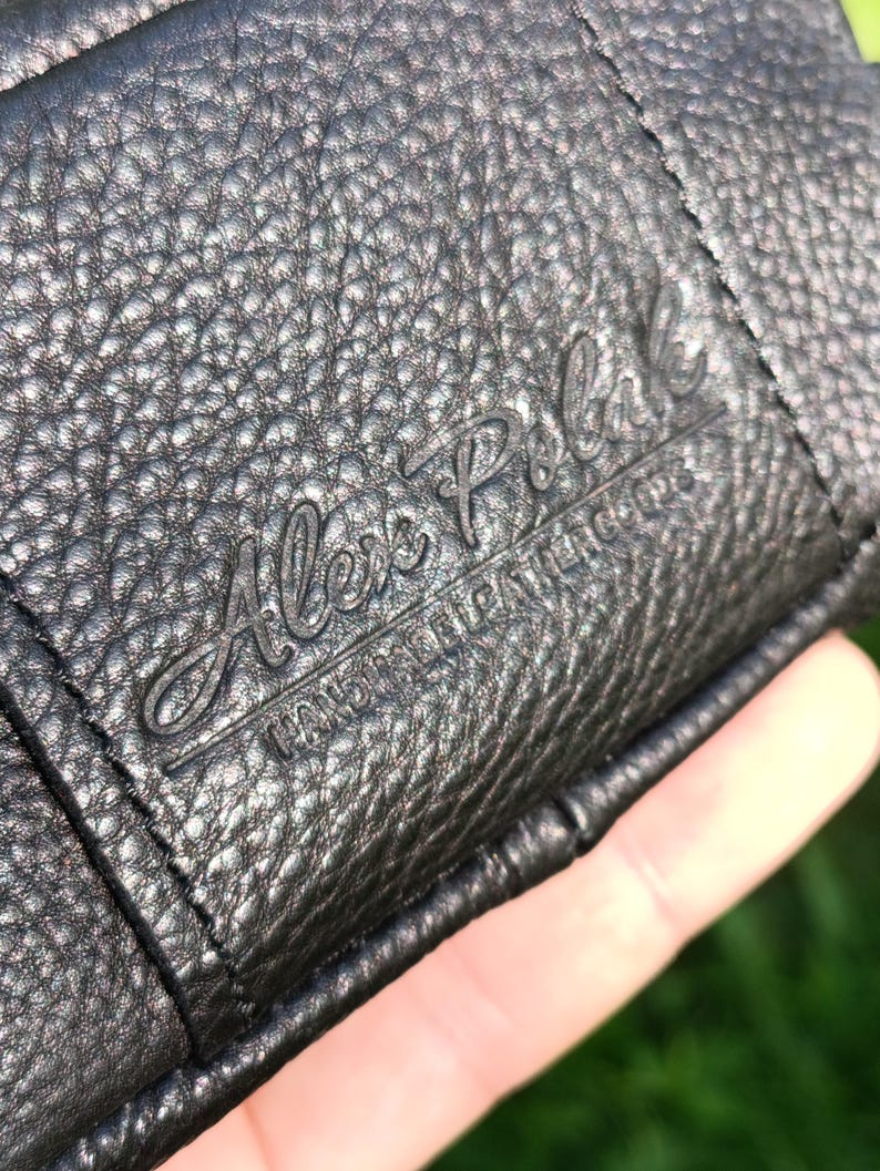 Puede incluir: Una cartera de cuero negro texturizado con el texto en relieve Alex Polak y las palabras HANDMADE LEATHER GOODS. La superficie tiene un patr&oacute;n de grano con costuras visibles a lo largo del borde del material de cuero doblado.