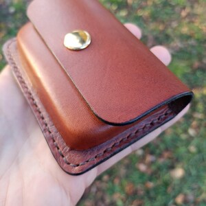 May include: A rectangular brown leather case featuring a smooth flap closure secured with a single polished gold-tone metal snap button. The edges of the case are reinforced with visible dark brown stitching along the perimeter.