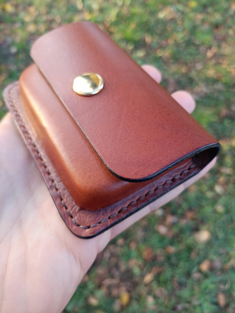 May include: A rectangular brown leather case featuring a smooth flap closure secured with a single polished gold-tone metal snap button. The edges of the case are reinforced with visible dark brown stitching along the perimeter.