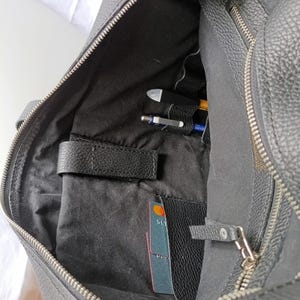 Puede incluir: Malet&iacute;n de cuero negro con cremallera abierta, que revela un interior con bolsillos y compartimentos. En el interior, hay bol&iacute;grafos, una tarjeta de cr&eacute;dito y un peque&ntilde;o bolsillo de cuero. El malet&iacute;n tiene una superficie texturizada y un asa.
