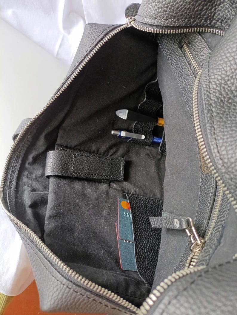 Puede incluir: Malet&iacute;n de cuero negro con cremallera abierta, que revela un interior con bolsillos y compartimentos. En el interior, hay bol&iacute;grafos, una tarjeta de cr&eacute;dito y un peque&ntilde;o bolsillo de cuero. El malet&iacute;n tiene una superficie texturizada y un asa.