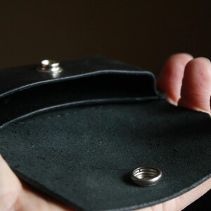 Puede incluir: Una cartera de cuero negro con cierre de presi&oacute;n y un detalle de anillo plateado. La cartera est&aacute; abierta y vac&iacute;a.