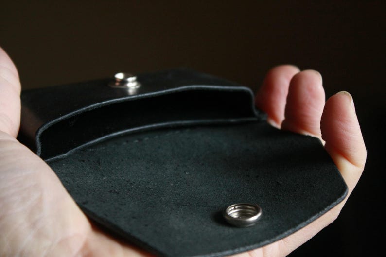 Puede incluir: Una cartera de cuero negro con cierre de presi&oacute;n y un detalle de anillo plateado. La cartera est&aacute; abierta y vac&iacute;a.