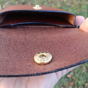 May include: A brown leather pouch featuring a gold-toned metal snap button closure. The interior of the pouch is visible, showing a smooth, dark brown leather lining with black finished edges along the opening of the compartment.