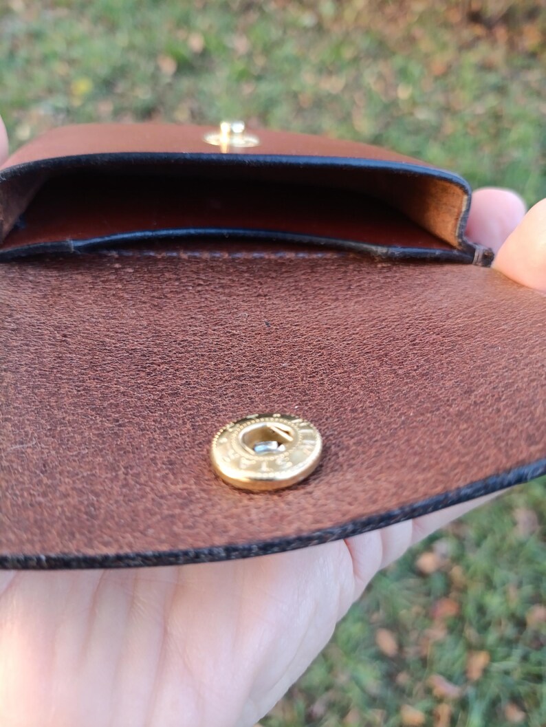 May include: A brown leather pouch featuring a gold-toned metal snap button closure. The interior of the pouch is visible, showing a smooth, dark brown leather lining with black finished edges along the opening of the compartment.