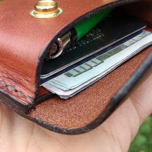 Puede incluir: Un tarjetero de cuero marr&oacute;n con dos botones de presi&oacute;n de metal dorado. El interior contiene un bol&iacute;grafo verde, una tarjeta de cr&eacute;dito negra y un fajo de billetes de cien d&oacute;lares visibles en el bolsillo abierto.