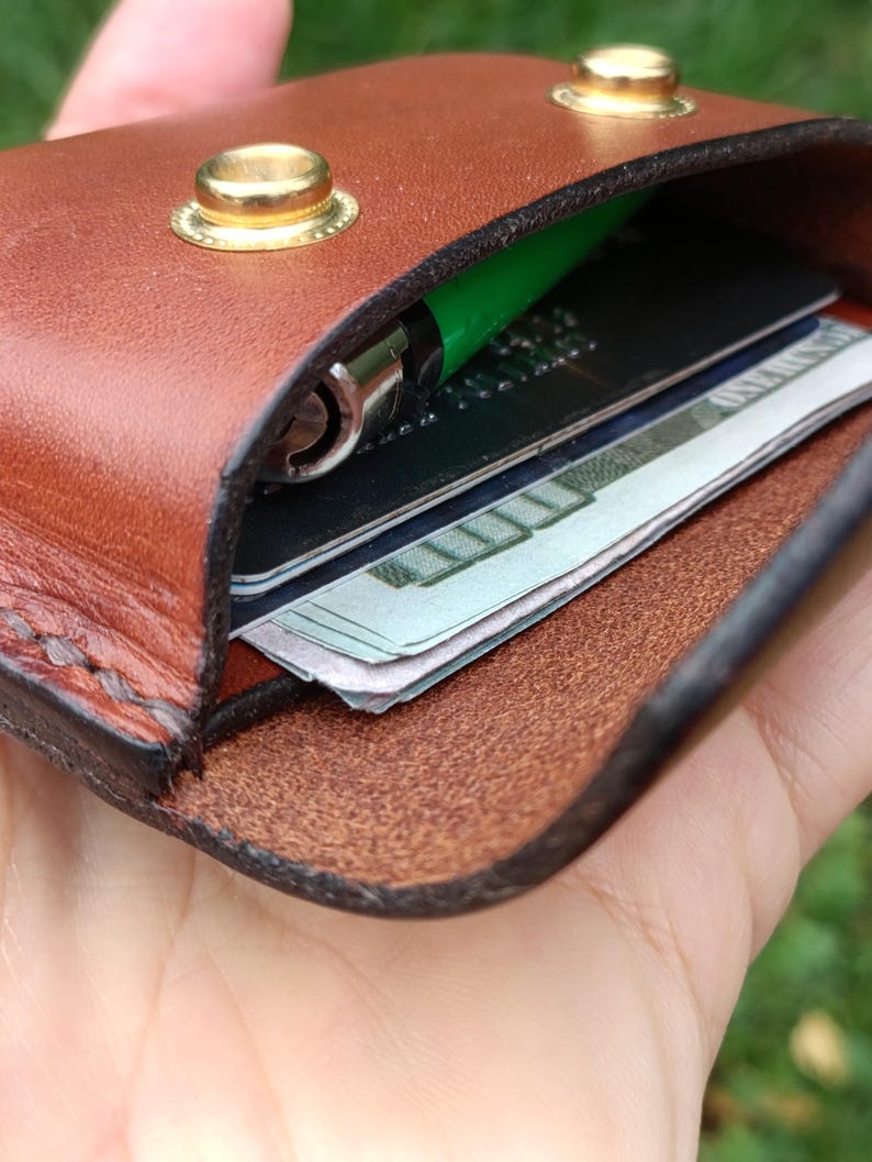 Puede incluir: Un tarjetero de cuero marr&oacute;n con dos botones de presi&oacute;n de metal dorado. El interior contiene un bol&iacute;grafo verde, una tarjeta de cr&eacute;dito negra y un fajo de billetes de cien d&oacute;lares visibles en el bolsillo abierto.