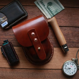 May include: A collection of leather goods and accessories on a wooden surface. Items include a brown leather pouch, a black wallet with a credit card, a pocket knife, a pocket watch, and a black lighter case. The pouch has copper rivets.