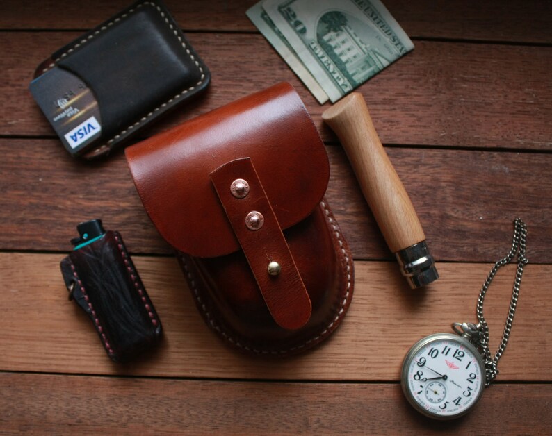 May include: A collection of leather goods and accessories on a wooden surface. Items include a brown leather pouch, a black wallet with a credit card, a pocket knife, a pocket watch, and a black lighter case. The pouch has copper rivets.